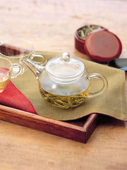 A Hario glass teapot filled with tea on a wooden tray with cloth and glass tea cups.